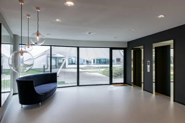 Moderne ontvangsthal met glazen hanglampen, een donkerblauwe bank, twee liften en grote ramen met uitzicht op een overkapt buitenterrein.